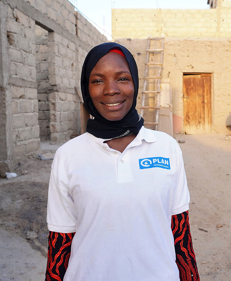 Eine junge Frau mit einem Plan T-Shirt steht in Timbuktu in einer Wohngegend zwischen Häusern.