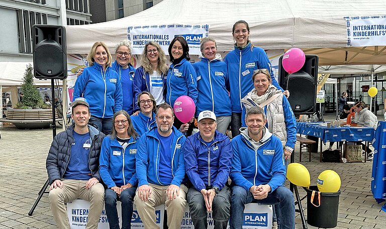 Mehrere Frauen und Männer in hellblauebn Jacken mit dem Plan-Logo sitzen vor einem weißen Festzelt