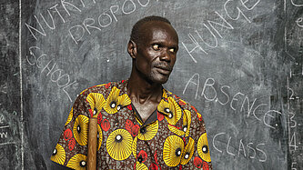Clement steht vor der Tafel. Darauf stehen die Worte "Hunger" "Nutrition" "Absence" und "Dropout" geschrieben.