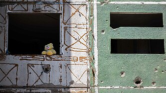 Eine Häuserfront mit Einschusslöchern, in einem Fenster sitzt ein Plüsch-Teddy