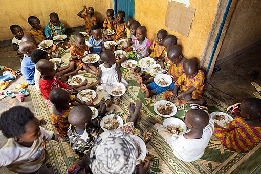 Eine Gruppe von Kindern sitzt auf dem Boden. Jedes Kind hat einen Teller mit Essen auf dem Schoß.