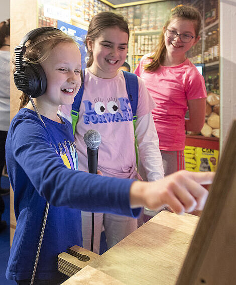 Kinder können in der interaktiven Ausstellung spielerisch lernen, was Entwicklungszusammenarbeit bedeutet Drei Mädchen sind bei einer Ausstellung und eine von ihnen bedient gerade einen aktiven Teil und trägt dafür Kopfhörer