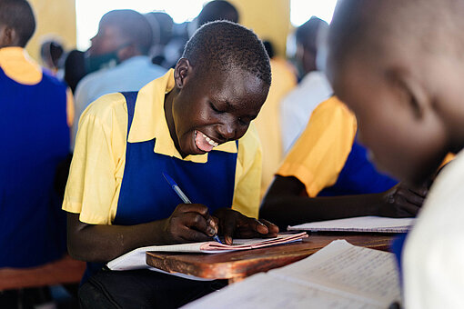 Die Kinder und Jugendlichen in Uganda können wieder in ihren Klassenräumen lernen – mit Unterstützung ihrer Lehrer:innen. Eine Schülerin in Schuluniform sitzt an ihrem Tisch und schreibt in ihr Heft.