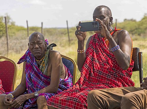 Maasai-Männer in roten Gewändern sitzen nebeneinander und einer hält ein Handy zum Fotografieren hoch.