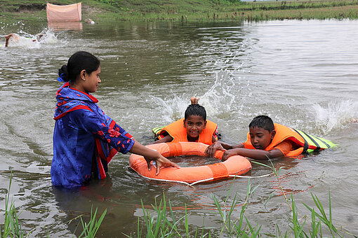 Bangladesch hat regelmäßig mit Überflutungen zu kämpfen. Daher ist Schwimmen eine lebenswichtige Kompetenz. Irin steht im Teich. Neben ihr schwimmen zwei Jungen in Schwimmwesten, die sich an einem Rettungsring festhalten.