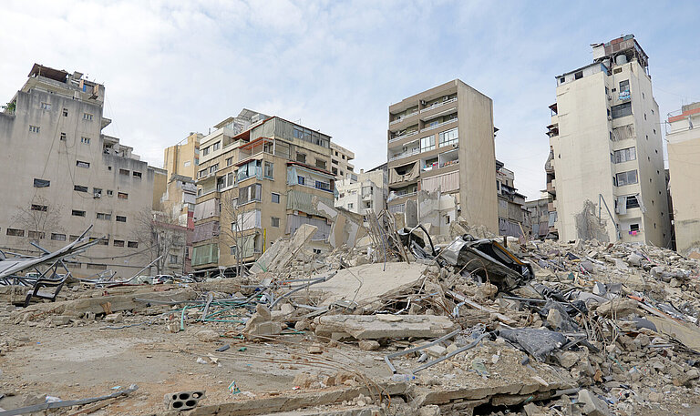 Eine zerstörte Wohngegend in der Stadt mit eingestürzten Gebäuden und Trümmern. Mehrere halb intakte Hochhäuser stehen im Hintergrund.