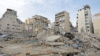 Eine zerstörte Wohngegend in der Stadt mit eingestürzten Gebäuden und Trümmern. Mehrere halb intakte Hochhäuser stehen im Hintergrund.