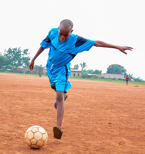 Junge in blauem Trikot und Flip-Flops visiert Ball an und holt zum Schuss aus 