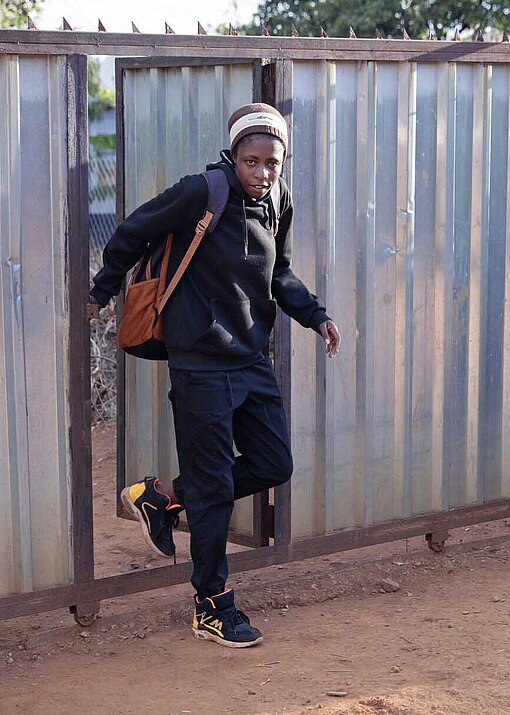 Eine Frau tritt aus einer Wellblechtür auf einen Sandweg