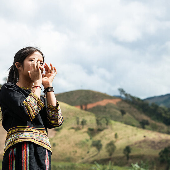 Ein Mädchen aus Vietnam steht draußen und hält beide Hände vor den Mund, als würde es etwas rufen
