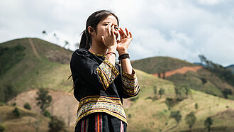 Ein Mädchen aus Vietnam steht draußen und hält beide Hände vor den Mund, als würde es etwas rufen