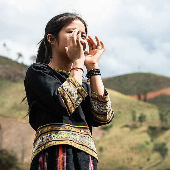 Ein Mädchen aus Vietnam steht draußen und hält beide Hände vor den Mund, als würde es etwas rufen