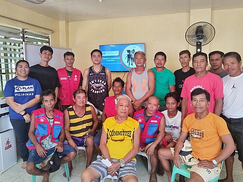 Eine Gruppe von etwa 15 Männern und Frauen steht gemeinsam in einem Raum. Sie posieren für das Foto und tragen verschiedene Kleidung. Links steht eine Frau mit Plan-Tshirt. Sie lächelt in die Kamera.