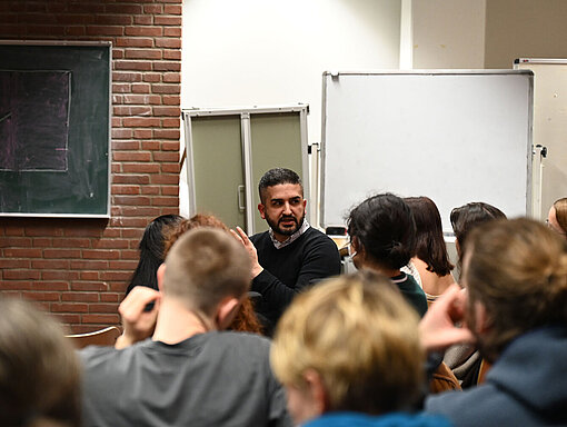 Gemeinsam mit dem Publikum wird diskutiert, was man von hier aus tun kann, um die Menschen im Iran zu unterstützen. Ein Mann sitzt im Publikum im Hörsaal und spricht mit den anderen.