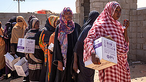 Frauen in Sudan stehen in einer Schlange und tragen Hilfspakete von Plan International.