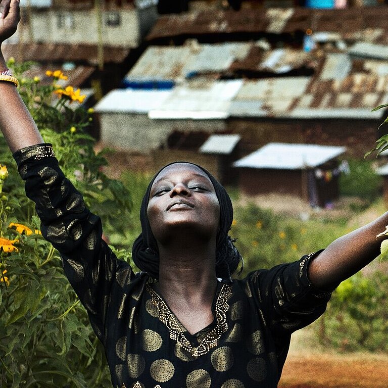 Eine Frau steht mit einer Blume in der Hand da, den Blick mit geschlossenen Augen zum Himmel gerichtet und die Arme von sich gestreckt. Im Hintergrund sieht man den Slum.