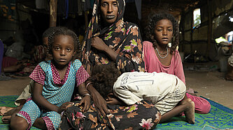 Geflüchtete Familie aus dem Sudan mit Mutter und Kindern, die in einer provisorischen Unterkunft sitzen.
