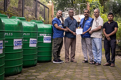 Fünf Männer stehen im Freien neben mehreren großen grünen Wassertanks, die mit dem Logo von Plan International beklebt sind. Zwei Männer halten gemeinsam ein Dokument in den Händen. 