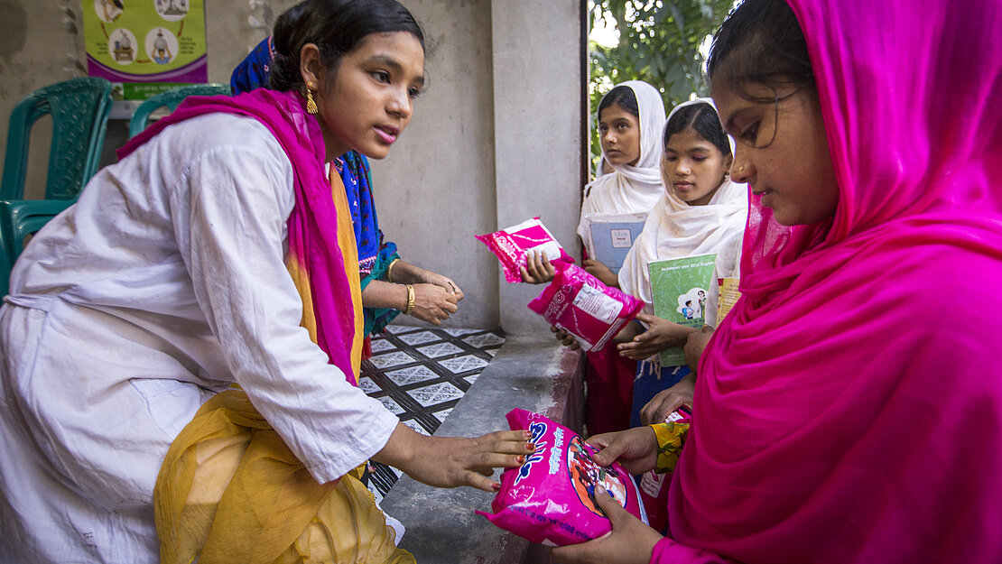 Ein Mädchen verkauft Bindenpackungen aus einem offenen Bereich der SaniMart-Produktionsstätte an drei andere Mädchen