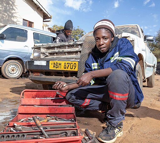 Eine junge Frau im Overall hockt in einer Kfz-Werkstatt vor einer Kiste mit Werkzeug