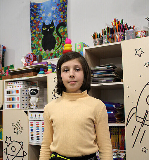 Der Kinderschutzraum ist für Helga zum Zufluchtsort geworden Ein junges Mädchen steht aufrecht vor einer Regalwand und lächelt leicht