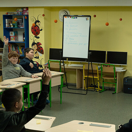 Eine Lehrerin steht vor einer kleinen Schulklasse in einem Klassenzimmer ohne Fenster