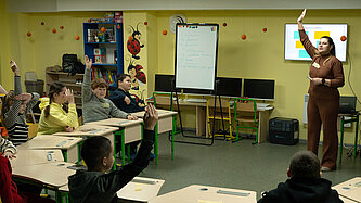 Eine Lehrerin steht vor einer kleinen Schulklasse in einem Klassenzimmer ohne Fenster
