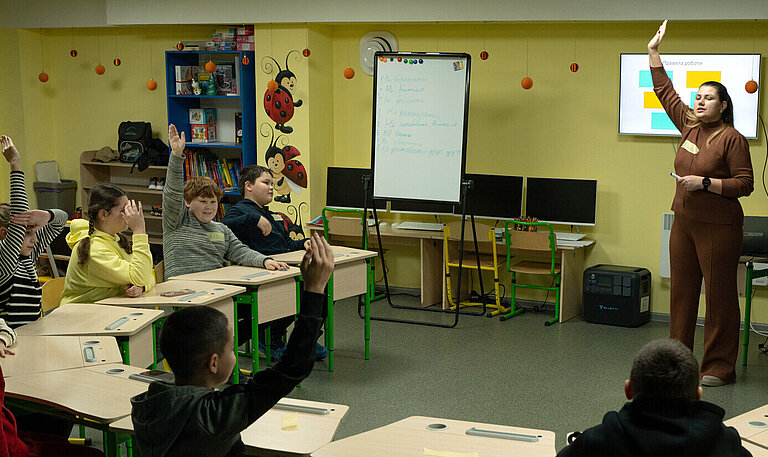 Eine Lehrerin steht vor einer kleinen Schulklasse in einem Klassenzimmer ohne Fenster