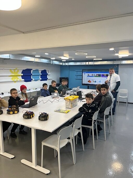 Mehrere Jugendliche sitzen an einem Tisch, auf dem Tisch stehen kleine Roboter-Autos, im Hintergrund ist ein großer Bildschirm. Der Raum hat keine Fenster.