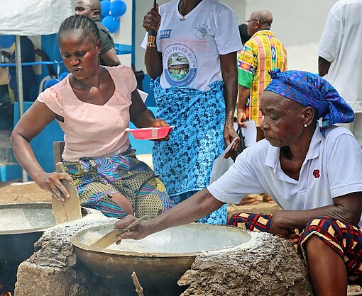 Mit hölzernen Keilen bereiten Frauen Fufu zu, ein nahrhafter Brei aus Maniok Zwei Frauen sitzen vor flachen Schalen, unter denen Feuer brennt, und rühren mit einem Holzkeil darin