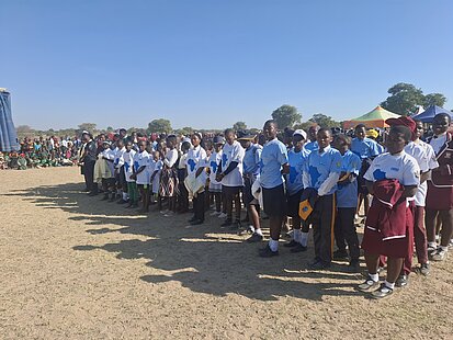 Eine Gruppe von Schulkindern steht in Reihen, einige tragen weiße T-Shirts und andere rote Schuluniformen. Die Kinder stehen unter freiem Himmel und schauen in die gleiche Richtung.