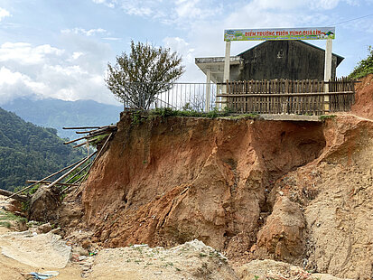 Eine Dorfschule steht auf einem durch Erosion gefährdeten Hang. Der Boden ist stark abgetragen, sodass Teile der Schule samt Zaun nahe am Abgrund stehen. Im Hintergrund sind grüne Berge und ein bewölkter Himmel zu sehen.