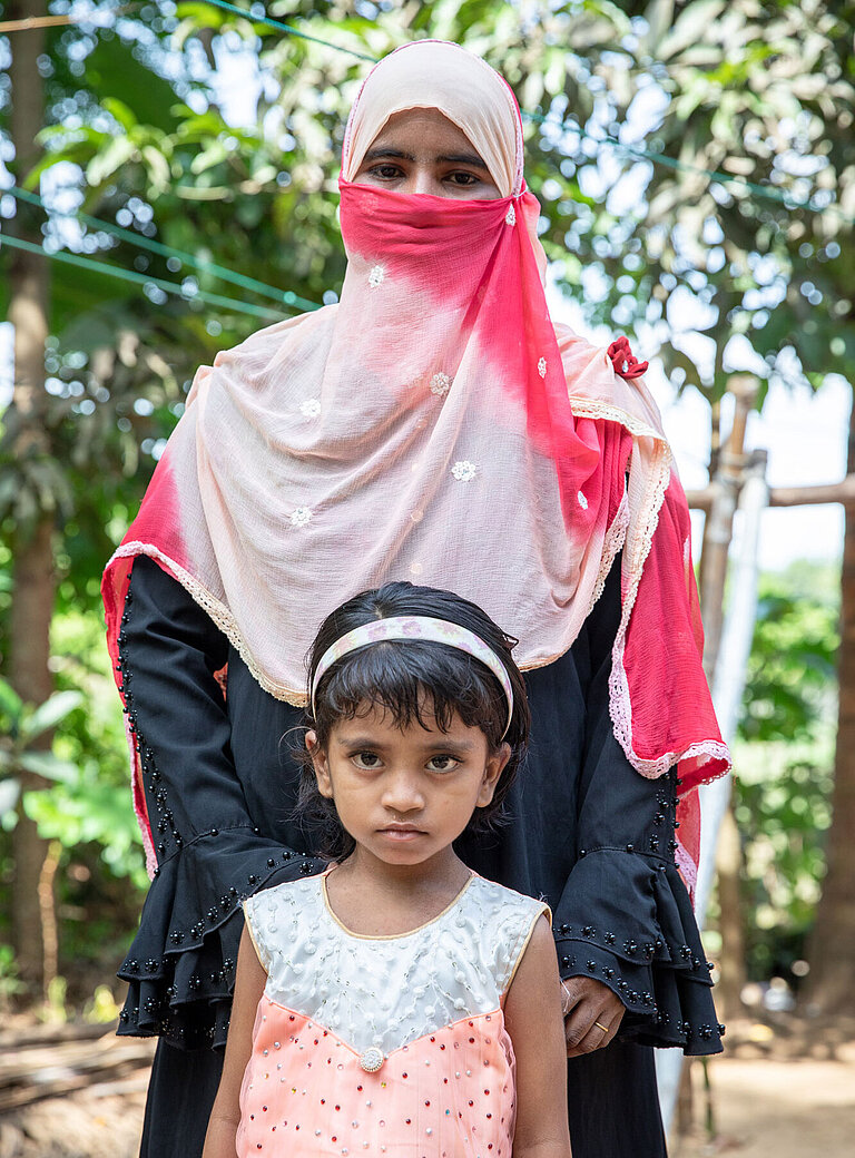 Eine Mutter steht hinter ihrer Tochter und hat ihr die Hände auf die Schultern gelegt. Die Mutter ist voll verschleiert und das Mädchen trägt ein rosa Kleid.