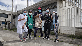 Fünf junge Erwachsene mit Masken posieren vor einer Bäckerei in Ecuador. Sie tragen Arbeitskleidung und zeigen Gesten der Zuversicht und Gemeinschaft.