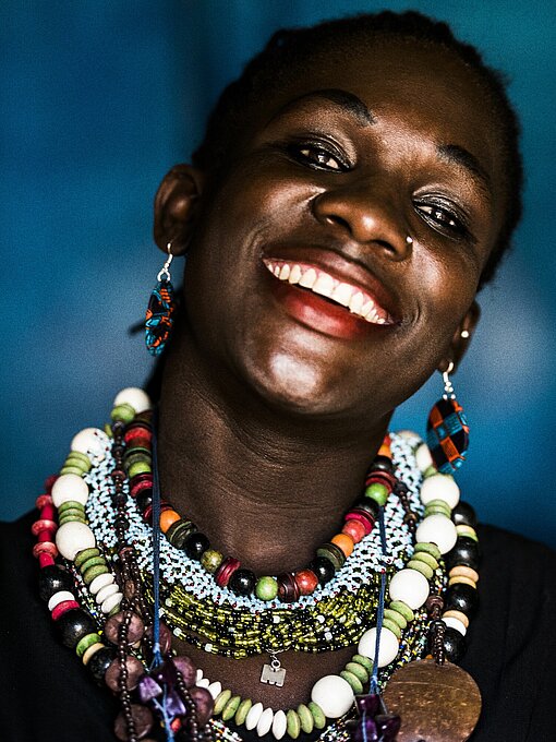 Marueen aus Kibera Portrait einer Schwarzen Frau, die den Kopf leicht seitlich gelehnt hat und lacht. Sie trägt viele verschiedene Ketten um den Hals.