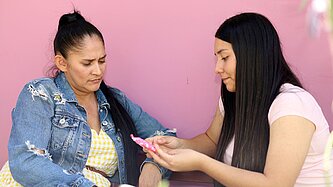 Mutter und Tochter sprechen miteinander. Sie sitzen vor einer pinken Wand, sitzen nebeneinander und sind in ein ernsten Gespräch vertieft