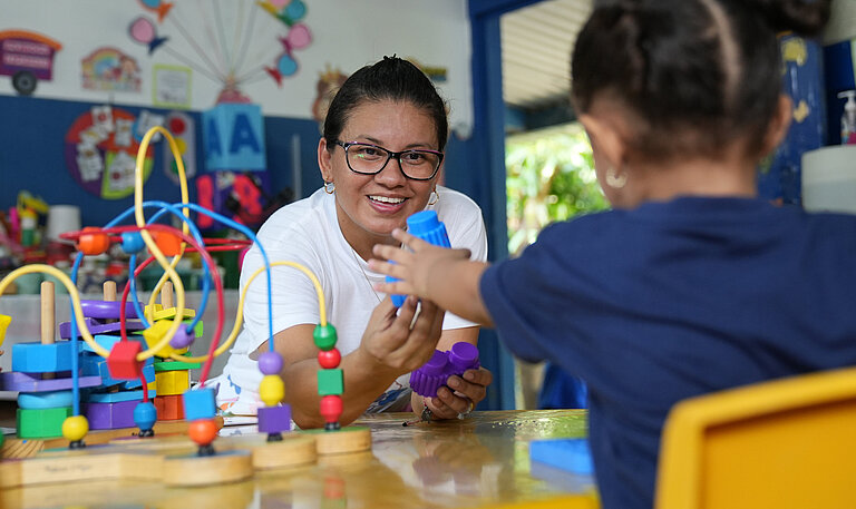 Eine Frau reicht einen blauen Bauklotz an ein junges Mädchen.