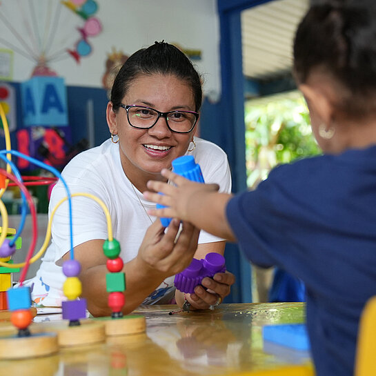 Eine Frau reicht einen blauen Bauklotz an ein junges Mädchen.