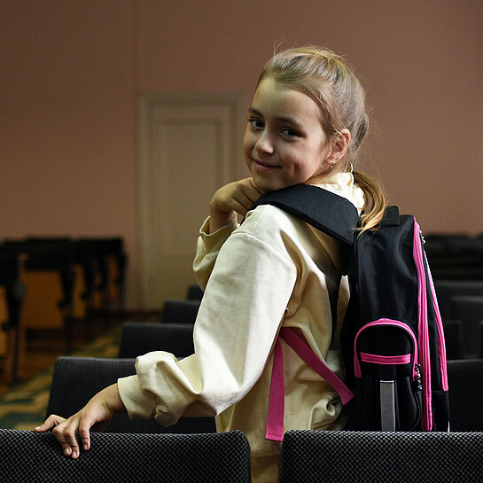 Ein Mädchen mit lila Rucksack schaut lächelnd über ihre Schulter.