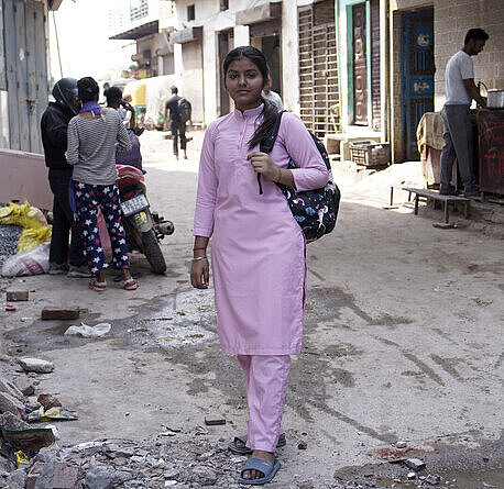 Neha ist auf dem Weg zu ihrer Arbeit in einer Klinik in Delhi Eine junge Frau in pinker Pflegerinnenkleidung und einem Rucksack über der Schulter geht zwischen Häuserreihen hindurch