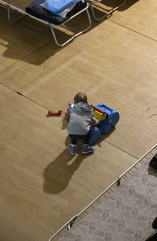 Kinder auf der Flucht benötigen Räume, in denen sie geschützt sind und Ablenkung bekommen. Ein Kind spielt auf dem Boden einer Turnhalle, die in ein provisorisches Lager für Geflüchtete umgebaut wurde.