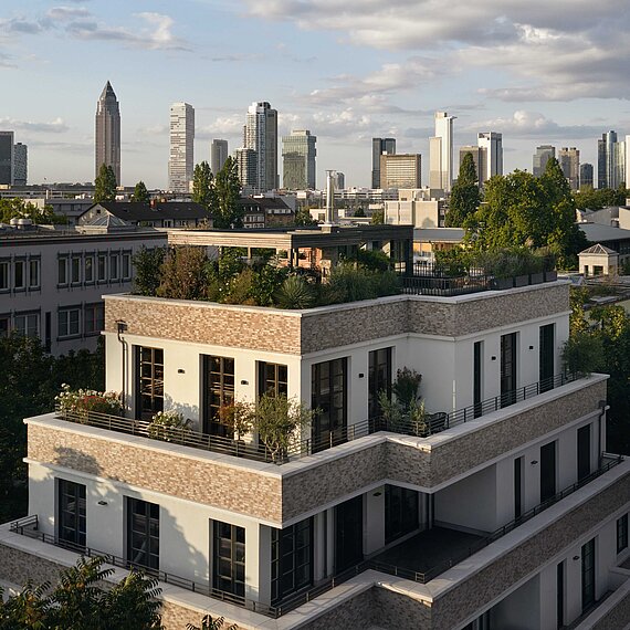 Ein modernes, mehrstöckiges Wohngebäude mit großzügigen, begrünten Terrassen steht im Vordergrund. Das Gebäude hat helle Fassaden und große Fenster. Dahinter sind zahlreiche Bäume sowie die Skyline einer Großstadt mit vielen Hochhäusern und markanten Gebäuden sichtbar. 