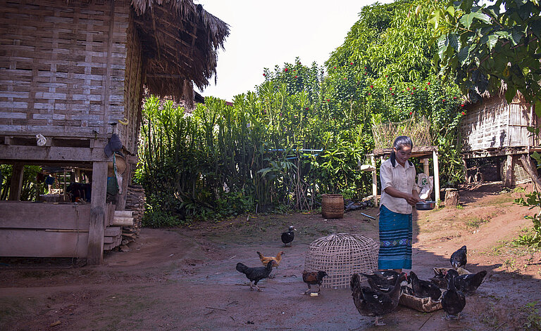 Eine Arbeiterin steht in ihrem Hof umgeben von Bananenpflanzen und füttert Hühner.