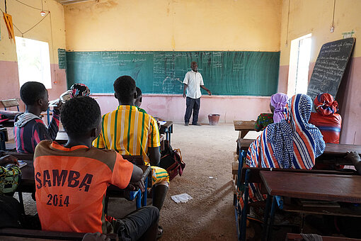 Bildung ist für viele Mädchen in Mali keine Selbstverständlichkeit. Ein Lehrer steht an einer Tafel und spricht mit Jugendlichen, die mit dem Rücken zur Kamera an ihren Schultischen sitzen.