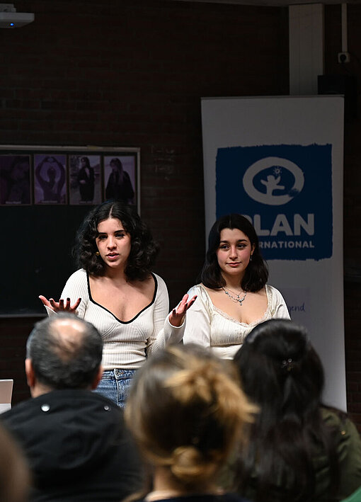 Zwei Mitglieder der Youth Advocates von Plan International Deutschland eröffnen einen Infoabend zu den Protesten im Iran. Zwei junge Frauen stehen in einem kleinen Hörsaal und sprechen zum Publikum.