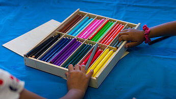 Kinderhände greifen nach einer großen Auswahl bunter Stifte in einer Holzbox auf einem blauen Tisch.