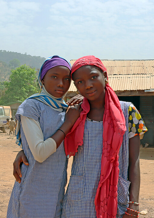 Zwei Mädchen Arm in Arm vor ihrem alten, baufälligen Schulgebäude und drei weißen Jeeps im Hintergrund