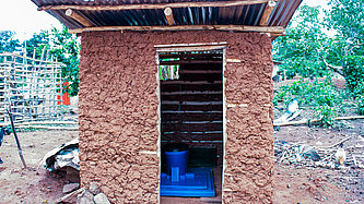Ein Toilettenhäuschen aus Lehm, im inneren sieht man eine Toilette aus blauem Kunststoff