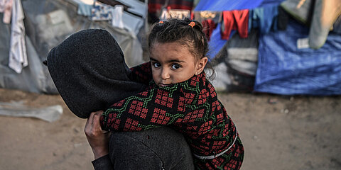 Ein Mädchen, das im Flüchtlingslager in Rafah im südlichen Gazastreifen lebt, wird von einer Person auf dem Rücken getragen. Sie schaut direkt in die Kamera. Im Hintergrund sind Zelte und Planen.