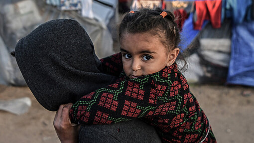 Ein Mädchen, das im Flüchtlingslager in Rafah im südlichen Gazastreifen lebt, wird von einer Person auf dem Rücken getragen. Sie schaut direkt in die Kamera. Im Hintergrund sind Zelte und Planen.